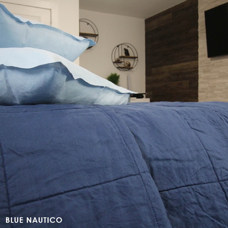 May include: A blue quilted bedspread with two light blue pillows on top. The bedspread is made of a soft, textured fabric. The pillows are also made of a soft, textured fabric. The bedspread is in a bedroom with a wooden wall and a television in the background.