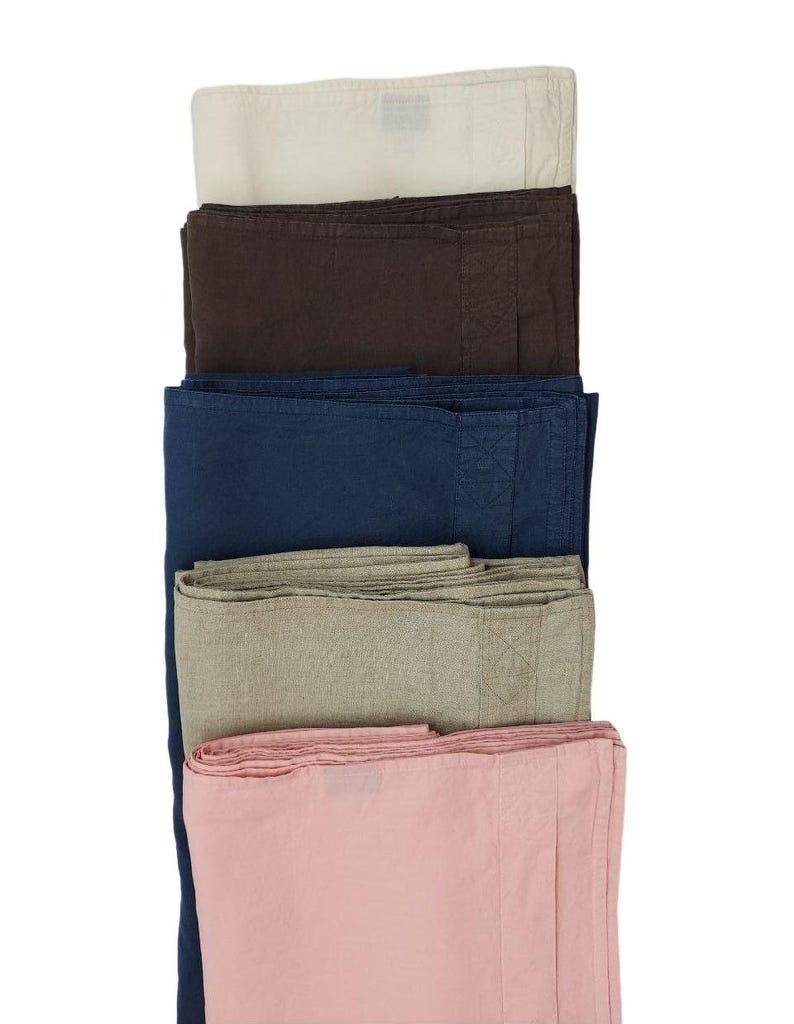 Op de afbeelding: Een stapel gevouwen linnen textiel in verschillende kleuren. De kleuren zijn gebroken wit, donkerbruin, marineblauw, lichtgroen en roze. De textiel is netjes opgevouwen, wat de textuur en de subtiele kleurvariaties benadrukt.