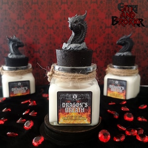 Peut inclure: Trois pots en verre avec des couvercles noirs et des sommets de tête de dragon. Les pots contiennent des bougies en cire de soja blanche avec des étiquettes qui indiquent "Dragon's Breath" et "Dougie Parfumge, Cire de Soja 100% Naturelle, www.Eitriandbrokkr.com". Les pots sont posés sur une surface noire avec des pierres précieuses rouges éparpillées autour d'eux.