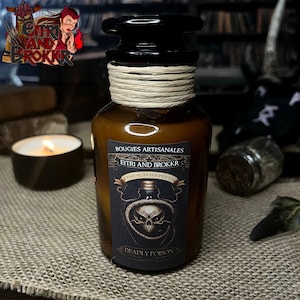 Pode incluir: Uma vela de vidro marrom em um frasco com um rótulo preto que diz "BOUGUIES ARTISANALES EITRI AND BROKKR VENENUM SERPENTIS DEADLY POISON". O rótulo apresenta um design de caveira e ossos cruzados.