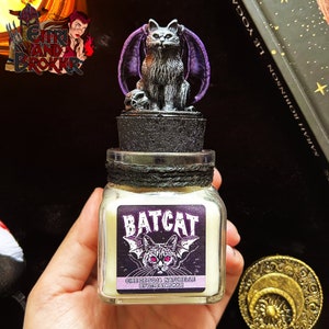 Peut inclure: Une petite bougie en verre dans un bocal avec une figurine de chat-chauve-souris noire et violette sur le dessus. La bougie est étiquetée "BATCAT" et "CIRE DE SOJA NATURELLE BY EITRI & BROKKR".