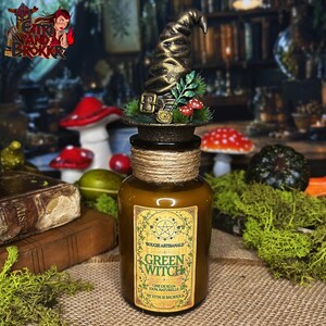 Peut inclure: Un pot à bougie en verre brun avec un col entouré de ficelle de jute, surmonté d'un chapeau pointu doré décoratif avec du feuillage vert et des champignons rouges. L'étiquette indique "BOUGIE ARTISANALE GREEN WITCH CIRE DE SOJA 100% NATURELLE BY EITRI & BROKKR".