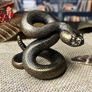 Pode incluir: Uma figurinha de metal dourada de uma cascavel enrolada em uma postura defensiva. A cobra tem uma superfície detalhada, texturizada e características realistas.