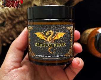 May include: A small, round jar with a black lid. The label features a golden dragon with outstretched wings and the words "DRAGON RIDER." The text "PAR EITRI & BROKKR - CIRE DE SOJA" is also visible. The jar is held in a hand.