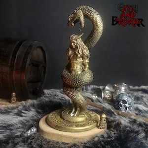 May include: A gold-colored figurine of a person wrapped around by a snake. The figurine is standing on a round base with runes inscribed on it. The base is made of wood and has a dark brown finish. The figurine is a representation of the Norse god, Jormungandr, the Midgard Serpent.