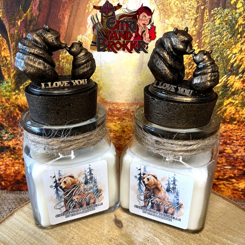 May include: Two glass jars with lids featuring a bear and cub sculpture. The jars contain soy wax candles with labels that read "Anne Elizabeth" and "Edward". The labels also include the text "Cire de Soja Naturelle" and "By Etti and Brokkr".