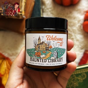Könnte beinhalten: Ein braunes Glas mit schwarzem Deckel, auf dem ein Etikett mit dem Text "Welcome to The Haunted Library" angebracht ist. Das Etikett zeigt eine gruselige Illustration eines Spukhauses, Kürbissen und Grabsteinen. Das Glas wird in einer Hand gehalten.
