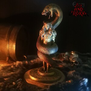 May include: A detailed figurine of a woman wrapped by a snake, with a gold base and runes. The figurine is painted in shades of blue, green, and gold. The text "G&Iacute;TRI AND BROKKR" is visible in the background.