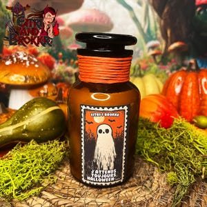 Peut inclure: Un bocal en verre brun avec une ficelle orange attachée autour du haut. Le bocal a une étiquette avec un fantôme et le texte "J'attends toujours Halloween..." Le bocal est posé sur un lit de mousse et de feuilles d'automne.