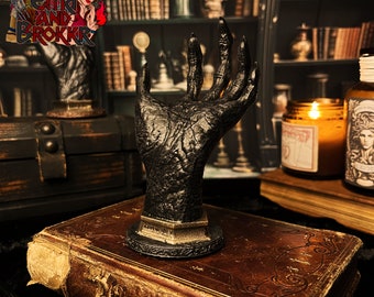 May include: A black resin hand sculpture with a detailed, textured surface. The hand is posed in a grasping gesture and is mounted on a black base with gold accents. The sculpture is displayed on a vintage book with a leather cover.