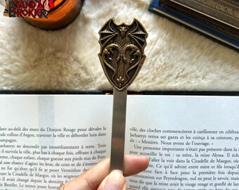 Può includere: Un segnalibro dorato con un drago che tiene una spada nelle sue grinfie. Il segnalibro ha la forma di uno scudo con una parte superiore appuntita e un bordo decorativo. Il drago è raffigurato con le ali spiegate e un'espressione feroce.