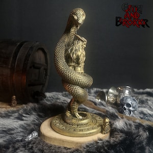 May include: A gold-colored metal sculpture of a snake wrapped around a human figure. The sculpture is on a circular base with runes inscribed around the edge. The sculpture is on a brown fur surface. The text "GITHRI AND BROKKR" is visible in the background.