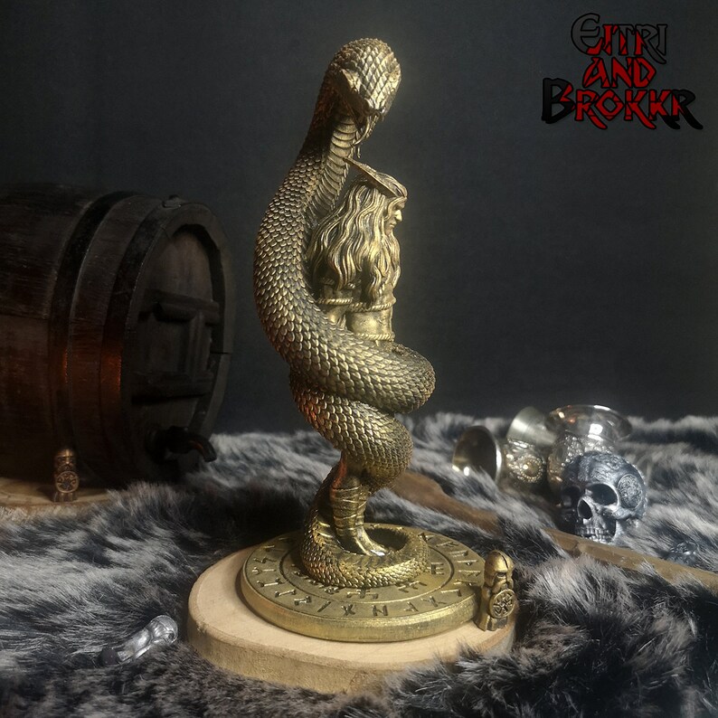 May include: A gold-colored metal sculpture of a snake wrapped around a human figure. The sculpture is on a circular base with runes inscribed around the edge. The sculpture is on a brown fur surface. The text "GITHRI AND BROKKR" is visible in the background.