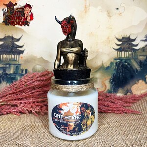 Op de afbeelding: Een glazen potkaars met een decoratieve Oni-Hime-thema topper. De topper heeft een rood gemaskerde figuur met hoorns en een kleine lantaarn. Het kaarslabel bevat Japanse tekst en de woorden "Princess Demon". De pot is omwikkeld met touw.