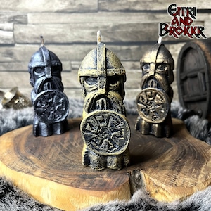 Könnte beinhalten: Drei dekorative Kerzen in Form von Wikingerkriegern, die jeweils einen Schild mit Runen halten. Die Kerzen sind in Schwarz, Gold und Silber.