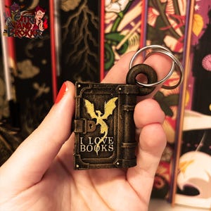 Peut inclure: Un porte-clés en forme de livre de couleur bronze avec un motif de dragon doré et les mots "I LOVE BOOKS". Le porte-clés a un anneau métallique et une boucle marron foncé. En arrière-plan, des dos de livres colorés.