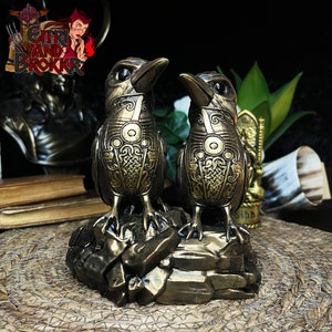 May include: Two golden raven figurines with intricate Celtic knotwork designs stand on a rocky base. The ravens have black eyes and beaks, and their wings are spread out.