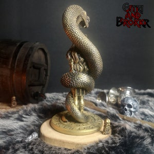 May include: A gold-colored metal statue of a woman with a snake wrapped around her. The statue is on a round wooden base with runes inscribed around the edge. The base is on a furry surface. The text "GITHRI AND BROKKR" is visible in the background.