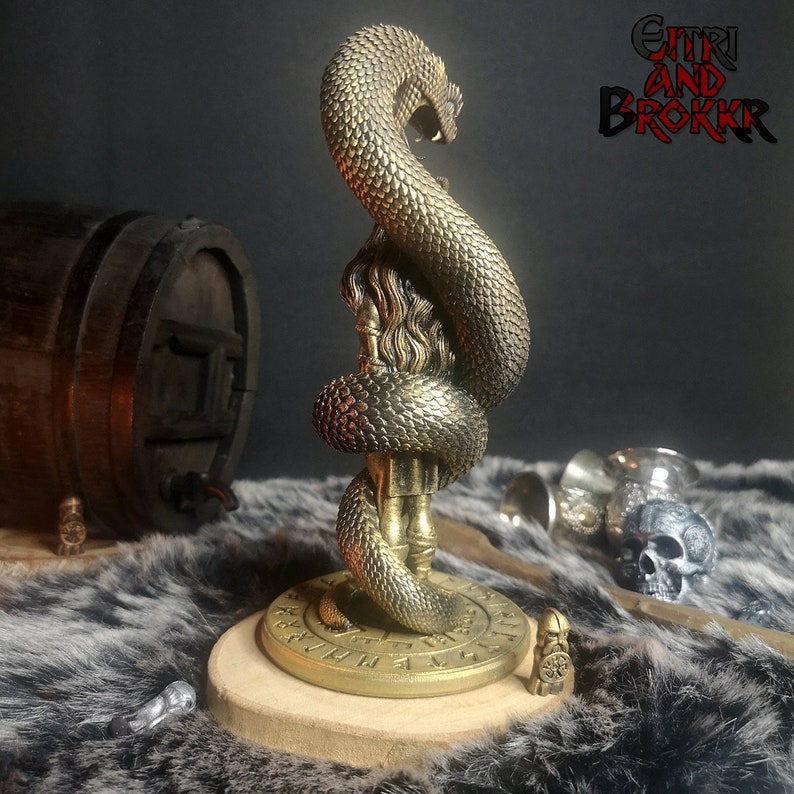 May include: A gold-colored metal statue of a woman with a snake wrapped around her. The statue is on a round wooden base with runes inscribed around the edge. The base is on a furry surface. The text "GITHRI AND BROKKR" is visible in the background.