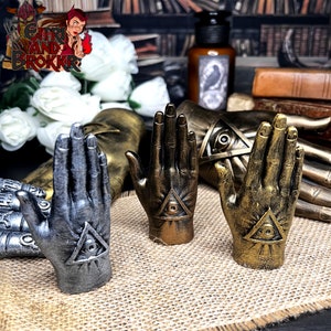 Op de afbeelding: Drie decoratieve handvormige beeldjes met een alziend oogontwerp. De handen zijn gemaakt van metaal en hebben een gouden, zilveren en bronzen afwerking.