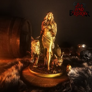 Może przedstawiać: Złota statua kobiety z długimi włosami i futrzaną peleryną, stojącej z dwoma wilkami u jej stóp. Statua stoi na drewnianej podstawie z okrągłym wzorem. Tekst "Eitri and Brokkr" jest widoczny w tle.