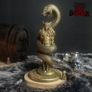 May include: A gold-colored figurine of a man with long hair and a beard, wrapped around by a snake. The figurine is standing on a round base with runes inscribed on it. The base is made of wood and has a dark brown color. The figurine is a representation of the Norse god, Jormungandr, the Midgard Serpent.