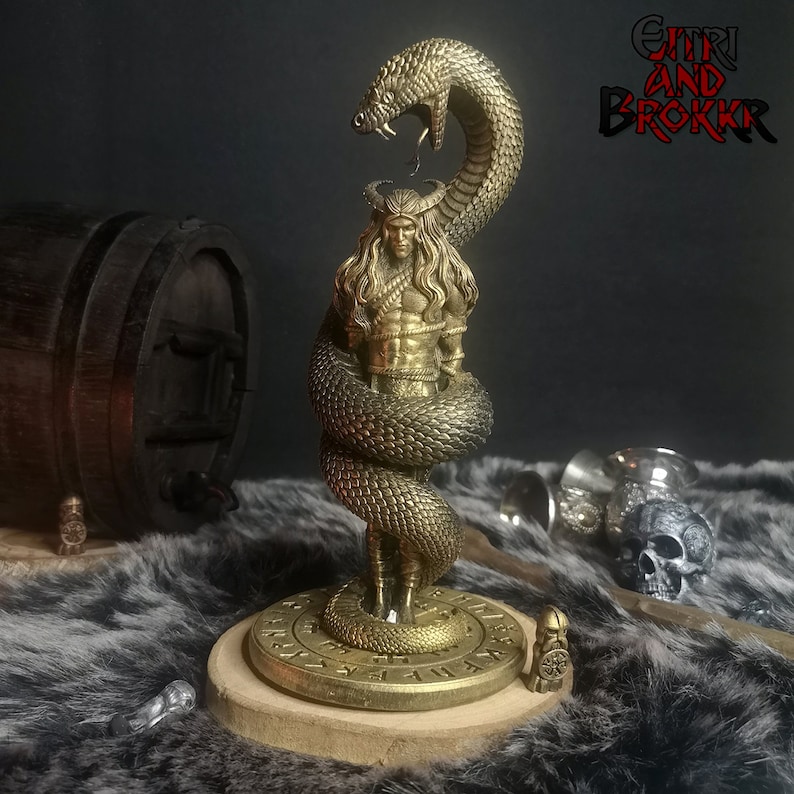 May include: A gold-colored figurine of a man with long hair and a beard, wrapped around by a snake. The figurine is standing on a round base with runes inscribed on it. The base is made of wood and has a dark brown color. The figurine is a representation of the Norse god, Jormungandr, the Midgard Serpent.