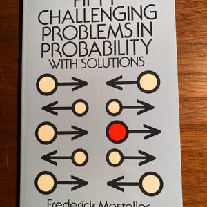 May include: A book cover with the title "Fifty Challenging Problems in Probability with Solutions" and the author's name "Frederick Mosteller". The cover features a geometric design with circles and arrows, with one circle highlighted in red.