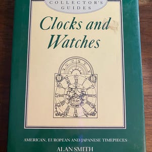 Può includere: Un libro con copertina rigida intitolato The Antique Collector's Guides Clocks and Watches di Alan Smith. La copertina è verde scuro con un pannello centrale color crema che presenta un disegno a linee nere di un orologio e American, European and Japanese Timepieces.