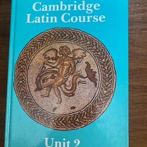 May include: A teal textbook titled "Cambridge Latin Course" with a mosaic illustration on the cover. The text "The North American Third Edition" is at the top, and "Unit 2" is at the bottom. The mosaic is brown and tan.