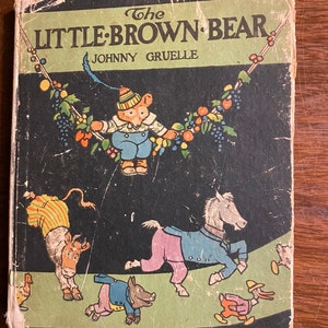 K&ouml;nnte beinhalten: Der Einband eines antiken Kinderbuches mit dem Titel "The Little Brown Bear" von Johnny Gruelle. Der Einband zeigt einen braunen B&auml;ren, der ein blau-wei&szlig; gestreiftes Hemd und eine rot-wei&szlig;e M&uuml;tze tr&auml;gt und an einer Rebe mit bunten Fr&uuml;chten und Beeren schwingt. Im Hintergrund sind weitere Tiere zu sehen, darunter eine Kuh, ein Pferd, ein Schwein und eine Ente.
