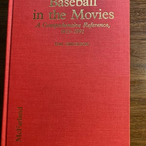 Puede incluir: Un libro de tapa dura rojo titulado "Baseball in the Movies" con letras doradas. El subtítulo del libro dice "A Comprehensive Reference, 1915-1991" y es de Hal Erickson. El nombre de la editorial, McFarland, está impreso verticalmente en el lomo.