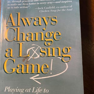 Può includere: Un libro blu con il titolo "Always Change a Losing Game!" di David B. Posen, MD. Il sottotitolo è "Playing at Life to Be the Best You Can Be".