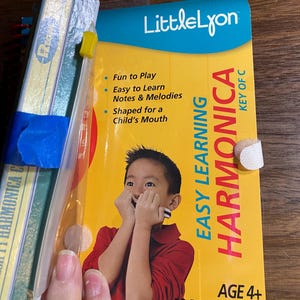 Può includere: Un kit di apprendimento per armonica "Little Lyon" giallo e blu. La copertina presenta un bambino che suona l'armonica, con il testo "Easy Learning Harmonica" e "Key of C". Il kit include un libro di 32 pagine ed è per bambini dai 4 anni in su.