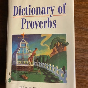 Könnte beinhalten: Ein weißes Buch mit einem blauen Streifen oben. Der Titel lautet "Dictionary of Proverbs" in blauer Schrift. Der Autor ist "David Pickering" in schwarzer Schrift. Das Cover zeigt eine Illustration eines Gewächshauses mit einem Hahn oben, einer Person auf einer Leiter und einem Vogel auf einem weißen Lattenzaun.