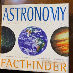 Op de afbeelding: Een blauw en wit boek getiteld "Astronomy Factfinder" met afbeeldingen van planeten en sterren. De omslag van het boek bevat de woorden "Jupiter, Kepler, Licht, Mercurius, Nevels, Oberon, Pandora, Quasar, Rigel, Saturnus, Titan 3, Schaduw, Viking, Witte dwerg, Xenon, Ziggoerats, Astronaut, Oerknal, Constellatie, Zonsverduistering, Vuurbal, Galaxie, Hubble, Ikrya-Seki, Jodrell Bank, Kitt Peak, Laika, Maan, Neptunus, Orion, Pluto, Quasar, Raket, Satelliet, Telescoop, Grote Beer, Venus, Gewichtloos".