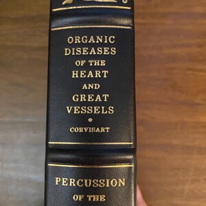 May include: A black book spine with gold lettering. The text reads "Organic Diseases of the Heart and Great Vessels" by Corvisart, and "Percussion of the Chest" by Auenbrugger. Gold floral embellishments are at the top of the spine.