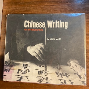 May include: A black and white book cover with the title "Chinese Writing: An Introduction" by Diane Wolff. The cover features a close-up of a hand writing Chinese characters with a brush.