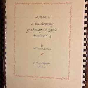 Op de afbeelding: Een beige boek met een titelpagina waarop staat "A Manual on the Acquiring of a Beautiful & Legible Handwriting" door William A. Bostick. Het boek is in 1977 uitgegeven door La Stampa Calligrafica in Detroit.