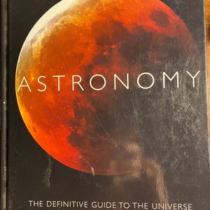 Op de afbeelding: Een zwart boek met de titel "Astronomie: De definitieve gids voor het universum" en een rood en oranje beeld van een planeet op de omslag. De tekst "Bevat de nieuwste foto's van de Hubble-telescoop en de Cassini-ruimtesonde" staat onder de titel.