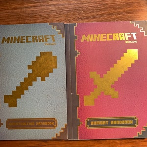 Puede incluir: Dos libros de guía de Minecraft, uno titulado "Construction Handbook" con una portada azul y el otro titulado "Combat Handbook" con una portada roja. Ambos libros presentan un gráfico de espada dorada y el logotipo de Mojang.