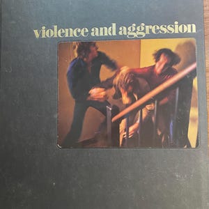 May include: A dark gray book titled "violence and aggression" in gold lettering. The book's cover features a photograph of a physical altercation. The image shows three people on a staircase, with one person appearing to be hitting another.