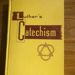 May include: A yellow hardcover book titled Luther's Catechism. The cover features dark brown rectangular accents behind the text and a geometric symbol consisting of three interlocking heart-shaped outlines centered below the title text.