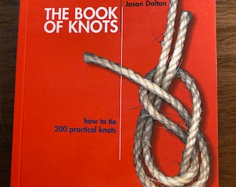 May include: A hardcover book titled The Book of Knots by Geoffrey Budworth and Jason Dalton, featuring a bright orange cover with a large, realistic illustration of a tied rope and the subtitle how to tie 200 practical knots.