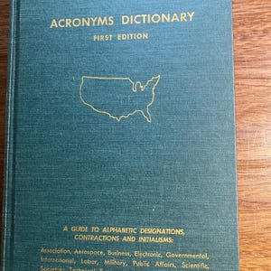 Peut inclure: Un livre bleu sarcelle intitulé "Acronyms Dictionary, First Edition". La couverture présente des lettres dorées et un contour doré des États-Unis. Le livre est un guide des désignations alphabétiques, des contractions et des initialismes.