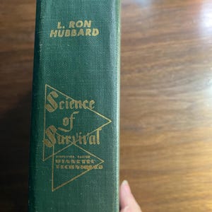 Op de afbeelding: Een vintage hardcover boek met een donkergroene kaft. De titel "Science of Survival" staat in gouden letters, samen met de naam van de auteur, L. Ron Hubbard. De rug van het boek bevat ook gouden tekst.