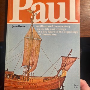 Puede incluir: Un libro titulado "Paul" de John Drane, un documental ilustrado sobre la vida y los escritos de una figura clave en los inicios del cristianismo. La portada presenta un barco con velas y una figura de proa de un ángel que sostiene una brújula.