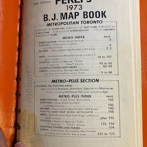 May include: An open vintage map book from 1973, titled "PERLY'S B.J. Map Book" for Metropolitan Toronto. The orange cover is visible, with text detailing the index, including hospitals, schools, and shopping centers. The book is open to page 1.