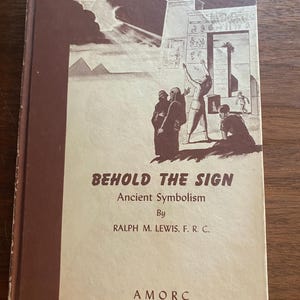 May include: A hardcover book titled Behold the Sign: Ancient Symbolism by Ralph M. Lewis, F.R.C. The cover features a brown and cream illustration of figures standing before an ancient temple with pyramids in the background and the text AMORC.