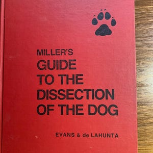 Può includere: Un libro rosso con il titolo "Miller's Guide to the Dissection of the Dog" di Evans & de Lahunta. Un'impronta di zampa nera è sulla copertina.